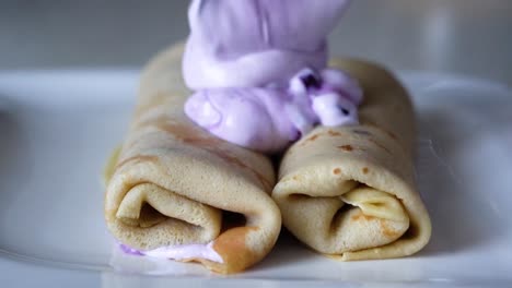 Two-rolled-French-pancakes-being-decorated-with-colorful-purple-blueberry-cream-on-a-white-plate