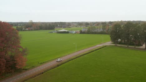 Drone-shot-following-silver,-grey-car-between-green-grass-fields-on-a-dutch-road,-street-from-above