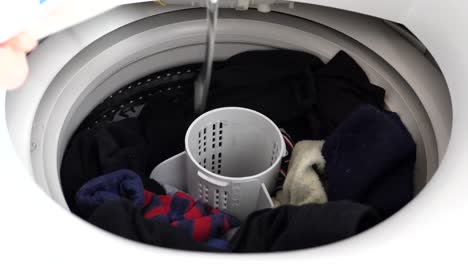 Hand-reaches-in-to-open-cap-on-white-washing-machine-appliance-to-pour-laundry-liquid-in-drum-of-dirty-load-with-colorful-socks-and-closing-lid-at-home