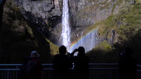 Schieben-Sie-Sich-In-Zeitlupe-In-Richtung-Silhouetten-Von-Menschen,-Die-Einen-Wunderschönen-Hohen-Wasserfall-Mit-Regenbogen-Betrachten
