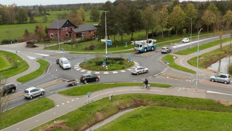 Drone-shot-of-dutch-roundabout-with-cars,-bikes,-trailors,-vans,-heavy-transportation-vehicles-passing-through,-while-surrounded-by-green-lush-grass
