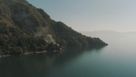 Drone-aerial-shot-of-Tzununa-bay,-smoke-coming-out-of-a-house,-Lake-Atitlan,-Guatemala