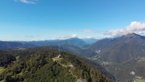 Communications-antenna-on-summit-of-vast-mountain-peak-valley-lush-green-hillside-aerial-pull-back