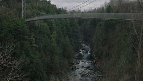 Estrecho-Río-Rocoso-Entre-Frondosos-árboles-Bajo-El-Puente-Colgante-En-Coaticook-En-Los-Municipios-Del-Este,-Quebec,-Canadá,-Tiro-Inclinado