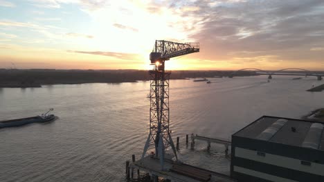 Aerial-sunset-view-of-a-small-container-terminal-crane-with-cargo-ships-passing-in-the-background
