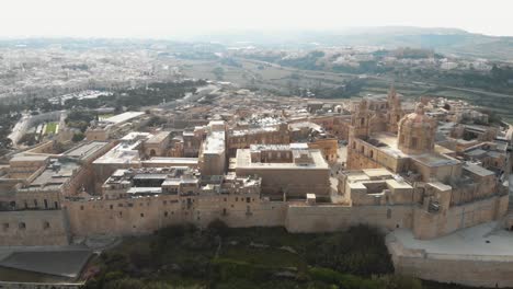 Mdina,-the-silent-city,-view-outside-city-walls-overlooking-the-cityscape-limestone-buildings-in-Malta---Ascending-panoramic-aerial-shot