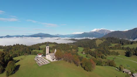 Drohne-Fliegt-In-Richtung-Der-Berühmten-Slowenischen-Kirche,-Grüne-Hügel-Und-Nebel-Im-Hintergrund