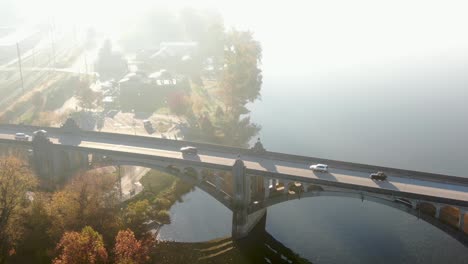 Cinematic-aerial-of-car-traffic-on-bridge-over-river-during-dramatic-morning-light-and-fog