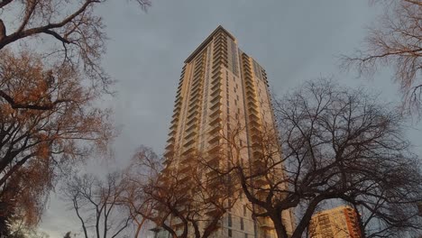 Stationary-camera-pan-out-upwards-of-clouds-moving-steady-around-a-modern-futuristic-luxury-apartment-building-next-to-vintage-towers-as-sun-sparkles-light-to-the-center-surrounding-by-bare-fall-trees