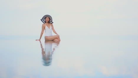 Asiatische-Frau-Mit-Weißem-Badeanzug-Und-Großem-Schwarz-weißem-Hut,-Die-Am-Rand-Des-Infinity-Pools-Sitzt-Und-Posiert