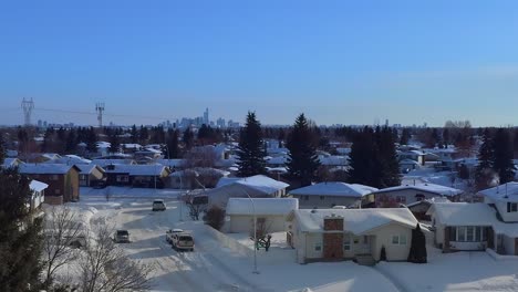 aerial-reveal-fly-over-snowed-in-dettached-lowrise-homes-snow-covered-streets-cars-reveal-by-tall-fraser-fir-tree-with-blue-skies-head-south-to-downtown-Edmonton-high-rise-skyscrappers-in-the-horizon