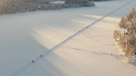 Vista-Aérea-Panorámica-Lateral-De-Un-Lago-Congelado-Y-Esquiadores-De-Fondo-En-Noruega-Con-Una-Puesta-De-Sol-Dorada-En-El-Fondo
