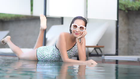 Attractive-Asian-woman-laying-on-edge-of-swimming-pool-kicking-feet-in-air-smiles-and-gives-sexy,-flirty-look-through-large-sunglasses