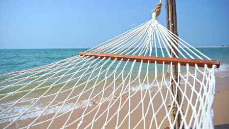 Close-up-of-a-rope-hammock-hanging-over-a-beach-and-incoming-waves