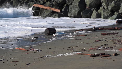 Foamy-Crashing-Waves-Against-Rocks-With-Wooden-Logs-On-Coastline