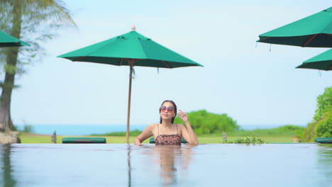 Asiatisches-Model,-Das-Mit-Sonnenbrille-Im-Swimmingpool-Steht-Und-Auf-Die-Tropische-Paradiesische-Landschaft-Des-Inselresorts-Mit-Meer-Und-Grünen-Sonnenschirmen-Im-Hintergrund-Blickt