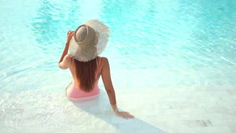 A-sexy-young-woman-in-a-bathing-suit-and-straw-hat,-with-her-back-to-the-camera,-sits-on-the-step-of-a-large-resort-pool