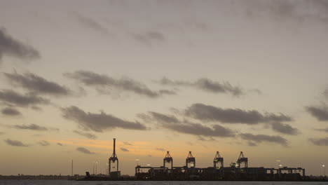 Tilting-shot-of-shipping-yard-in-the-distance-at-sunset-with-wooden-pylons-in-the-foreground