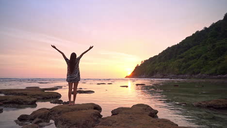 A-Woman-Standing-on-Rock-at-Tropical-Beach-and-Raising-Hands-on-Sunset-Sunlight