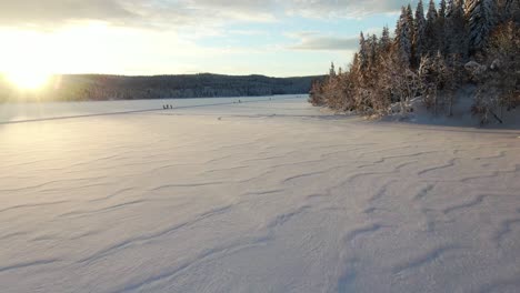 Vista-Aérea-De-Baja-Altitud-De-Un-Lago-Congelado-Y-Esquiadores-De-Fondo-En-Noruega-Con-Puesta-De-Sol-Dorada-Y-Paisaje-Invernal-En-El-Fondo