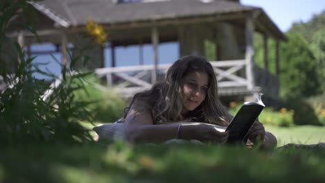 Junge-Lateinamerikanische-Frau-Lächelt-Und-Studiert-Lesebuch-Im-Freien-Auf-Dem-Gras-Mit-Vegetation-Und-Haushintergrund-Zeitlupe-60-Fps