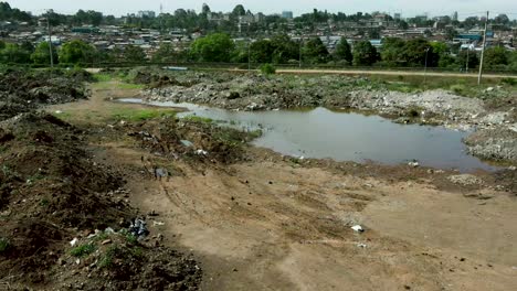 Dirty-dumping-site-with-stagnant-unhealthy-water,-rubbish-and-trash-separated-by-highway-and-sad-urban-slums-in-background