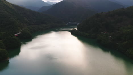 Vista-Pintoresca-De-Un-Río-Sereno-Rodeado-De-Montañas-Exuberantes-Con-Puente-Colgante-En-El-Lago-Okutama,-Japón