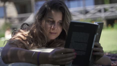Young-latin-female-interested-studying-reading-book-outdoors-on-the-grass-with-a-vegetation-and-house-background-Slow-Motion-60-fps