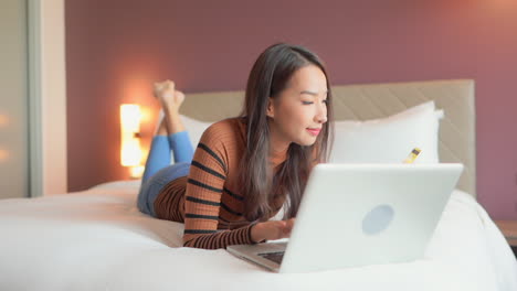 Beautiful-Asian-woman-lying-on-the-bed-with-her-laptop-in-front-of-her-and-types-in-the-credit-card-number-to-pay-for-food-delivery