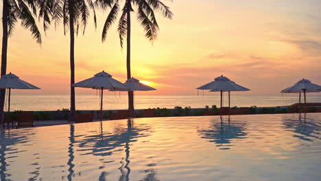 Perfekter-Sonnenuntergang-Mit-Roter-Sonne-Von-Der-Lounge-Des-Tropischen-Hotelresorts-In-Der-Nähe-Des-Infinity-Pools,-Sonnenschirmen-Und-Kokospalmen-Im-Vordergrund