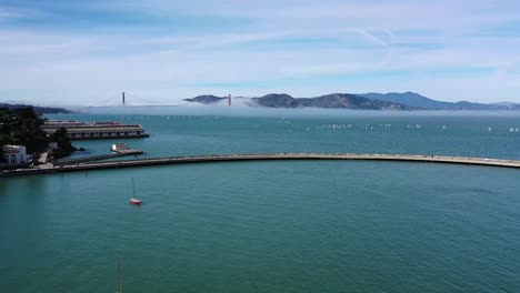Drone-flyover-of-Aquatic-Park-Pier-in-San-Francisco-with-the-Golden-Gate-bridge-in-the-background-shrouded-with-light-fog-on-a-sunny-day