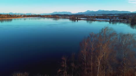 4K-UHD-aerial-drone-flight-moving-backwards-above-an-icy-frozen-lake-and-tree-tops-in-Bavaria-close-to-the-Alps-in-winter-in-Germany-near-Austria
