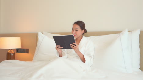 Smiling-pretty-Asian-woman-lying-on-the-hotel-white-bed-wearing-a-white-dressing-gown-and-reading-a-book-at-night-time