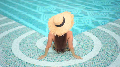 Back-of-glamorous-female-model-in-a-swimsuit-and-summer-hat-sitting-in-a-shallow-swimming-pool-water,-full-frame-slow-motion