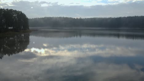 Aerial-shot-on-mystic-foggy-morning-over-the-calm-lake-hidden-in-the-forest-on-autumn-with-sky-reflects-on-a-flat-surface