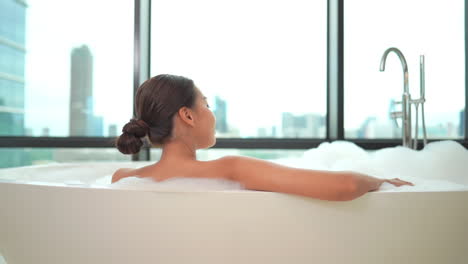 A-woman-sits-in-a-luxurious-bubble-bath-while-looking-out-at-a-modern-city-skyline
