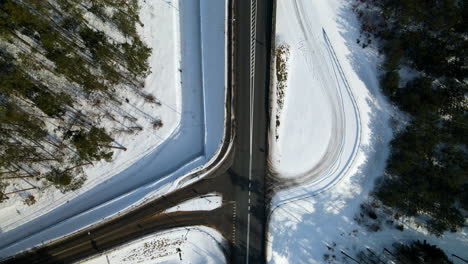 Drone-over-black-road-near-forest-during-snowy-winter,-moderate-traffic,-top-down,-Poland
