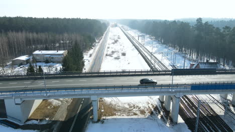 Cars-driving-over-a-viaduct-crossing-another-black-road-and-three-tracks-of-a-railway-near-a-station-between-the-trees-on-a-winter-landscape