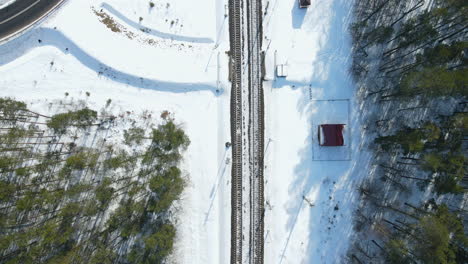 Aerial-top-down-of-empty-snow-covered-rail-tracks,-small-train-station-and-pine-trees-during-sunny-day-in-winter