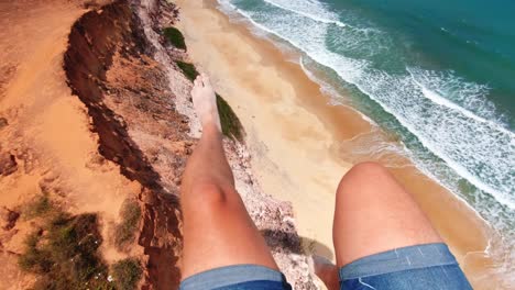 Legs-of-a-man-sitting-in-a-chair-of-a-paraglide-with-sandy-cliffs-and-beautiful-blue-ocean-water-below-in-the-tropical-city-of-Pipa-in-Rio-Grande-do-Norte,-Brazil-on-a-warm-sunny-summer-day