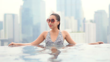 Junge-Asiatische-Frau-Mit-Sonnenbrille-Im-Städtischen-Pool-Auf-Dem-Dach-Und-Wolkenkratzern-Im-Hintergrund
