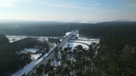 Aerial-flyover-rural-intersection-surrounded-by-large-forest-trees-during-snowy-winter