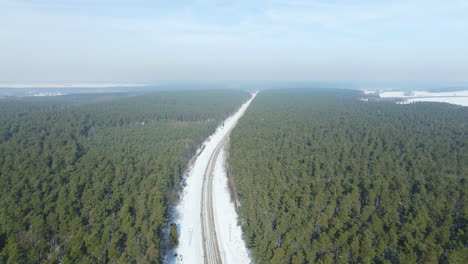 Beautiful-snow-covered-railway-road-through-the-evergreen-coniferous-forest-in-Rakowice,-Poland---aerial-slow-fly-over-shot