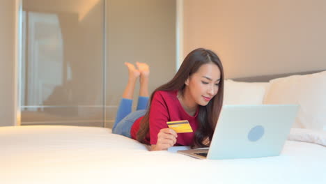 Eine-Junge-Asiatische-Frau,-Die-Online-Mit-Kreditkarte-Und-Laptop-computer-Im-Bett-Des-Einfamilienhauses-Einkauft