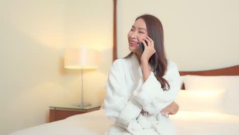 Glückliche-Asiatische-Frau-Im-Bademantel-Auf-Dem-Smartphone,-Die-Nachtanruf-In-Einem-Schlafzimmer-Chattet