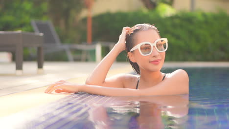 A-close-up-young-attractive-woman-wearing-sunglasses-in-a-swimming-pool-leans-on-the-edge-of-the-pool-turns-to-look-around