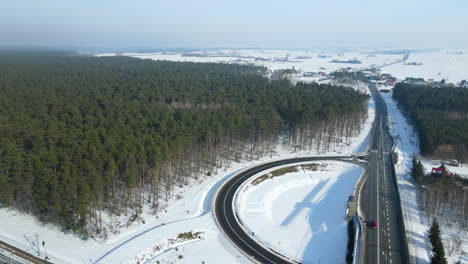 Vehicles-Traveling-On-The-Highway-And-Curve-Road-With-Green-Forest-On-The-Snowy-Field-In-Rakowice,-Krakow,-Poland