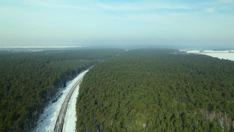 Snowy-Country-Road-Through-Lush-Forest-Landscape-On-A-Sunny-Winter-Day-In-Rakowice,-Poland---Aerial-Drone-Shot
