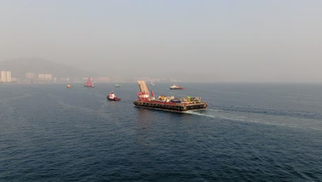 Barcaza-Cargada-Con-Camiones-Hormigonera-Tirada-Al-Puerto-Por-Un-Remolcador-En-La-Bahía-De-Hong-Kong,-Vista-Aérea