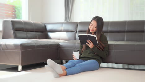 A-pretty-young-woman-sitting-on-the-floor-holding-a-cup-of-coffee-while-she-reads-a-book
